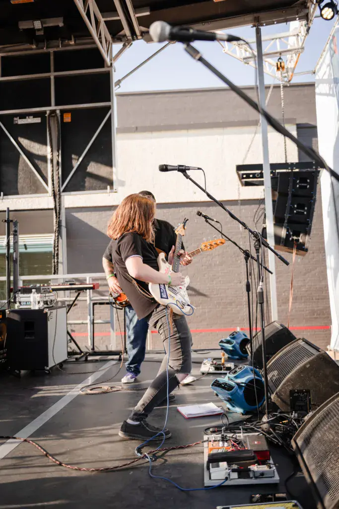 A musician with reddish hair plays electric guitar on stage under bright sunlight at an outdoor music festival.