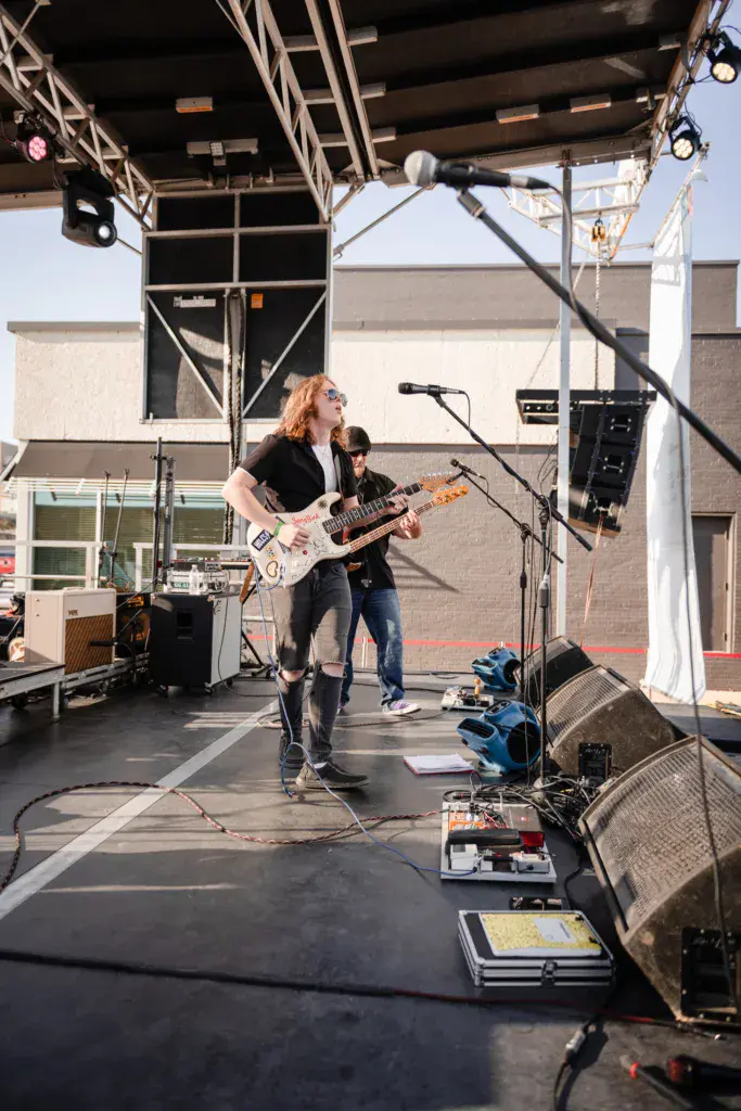 A guitarist with reddish hair performs on stage under a metal truss structure at an outdoor music festival.