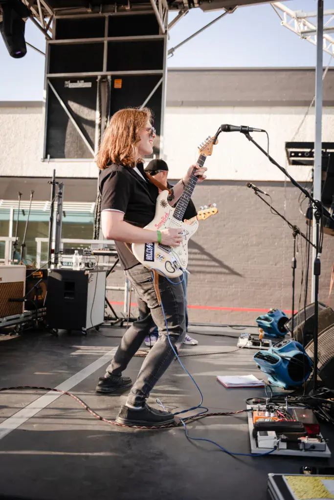 A guitarist with reddish hair performs on an outdoor stage, playing a white electric guitar at the microphone.