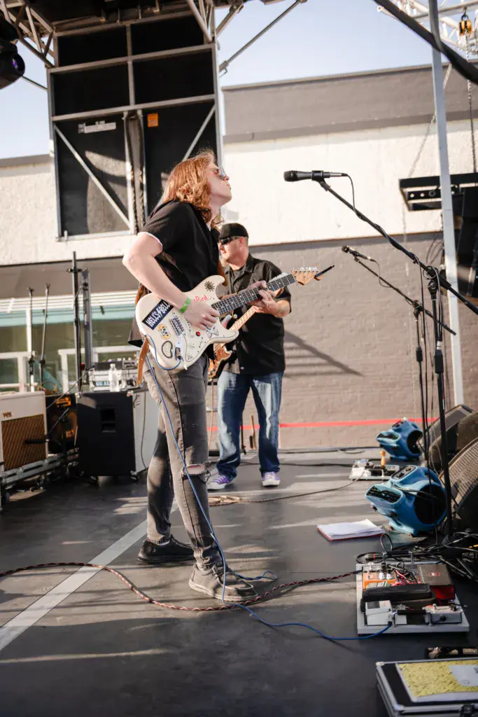 A musician with reddish hair plays electric guitar on stage while another band member plays guitar in the background.