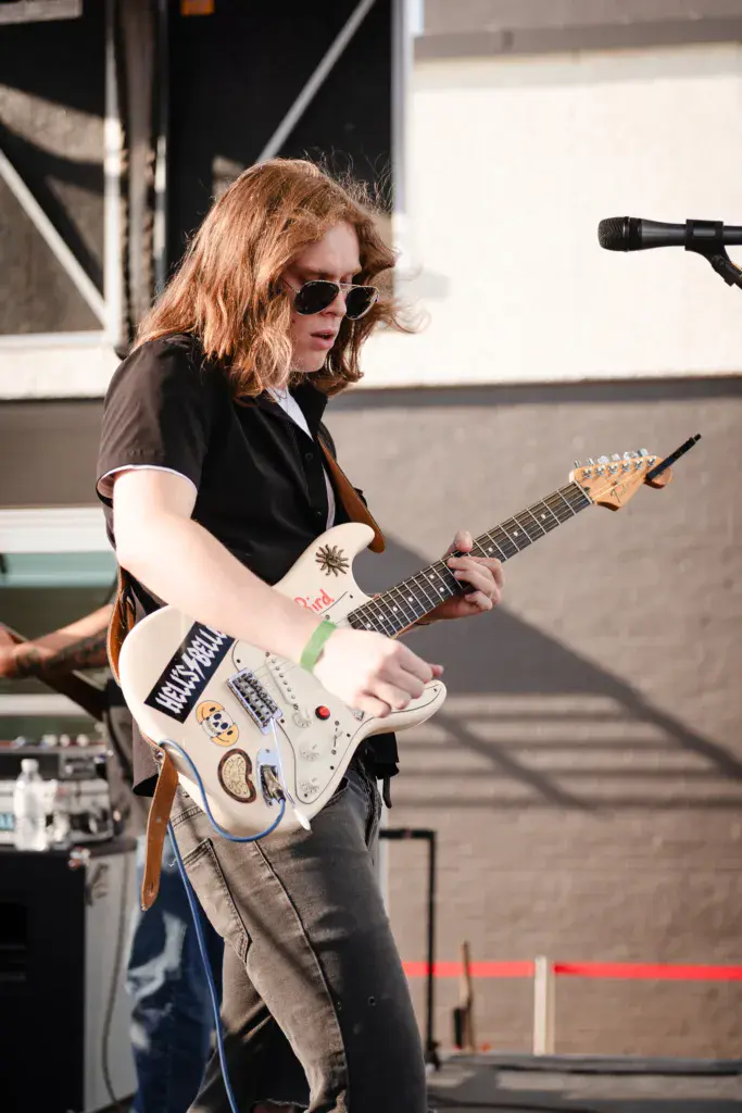 A guitarist with reddish-brown hair wearing sunglasses and a black shirt plays a white electric guitar on stage with a microphone visible nearby.