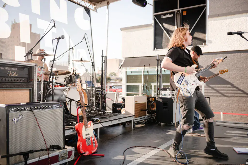 A band performs on an outdoor stage with a guitarist in the foreground and drummer on a raised platform.