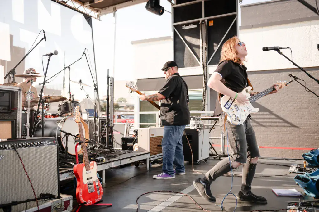 A band performs on an outdoor stage at a music festival, with a guitarist, bassist, and drummer playing together.