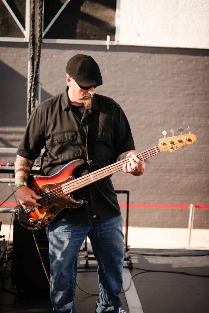 Bass guitarist in black cap and shirt performing on stage at an outdoor music festival.