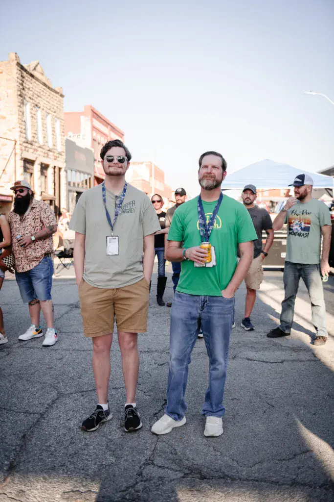 Festival attendees wearing lanyards stand on a street with historic brick buildings and white tents in the background.