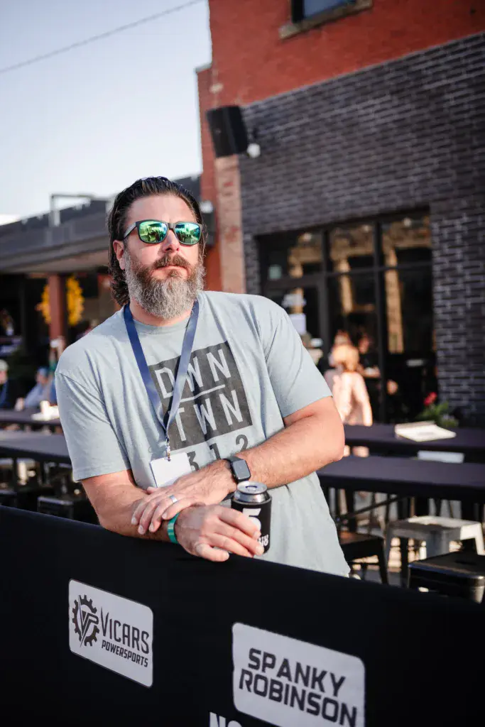 A bearded man wearing sunglasses and a festival lanyard stands at an outdoor venue holding a microphone.