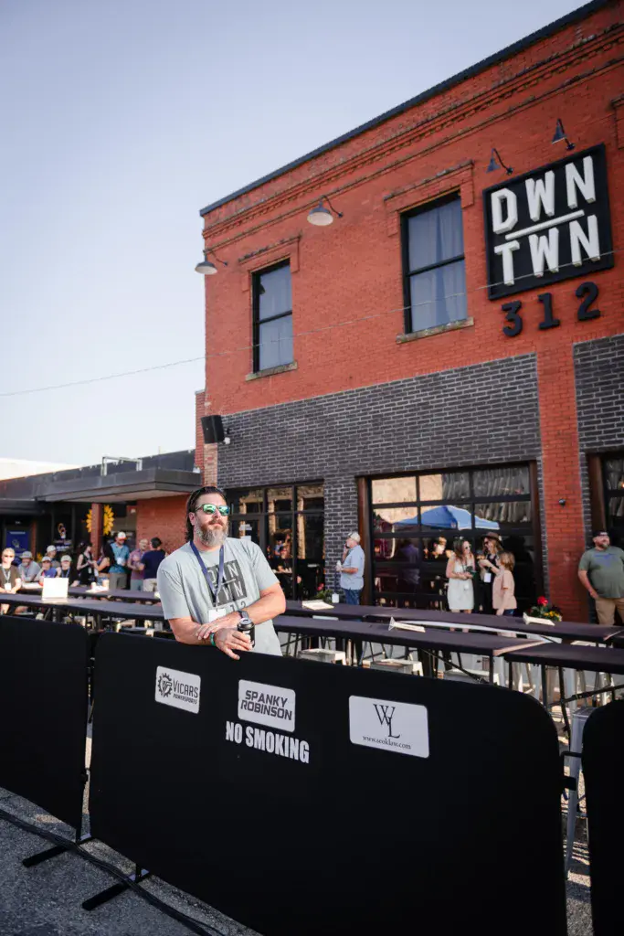 A performer stands at a DJ booth in front of a red brick downtown building with festival attendees in the background.