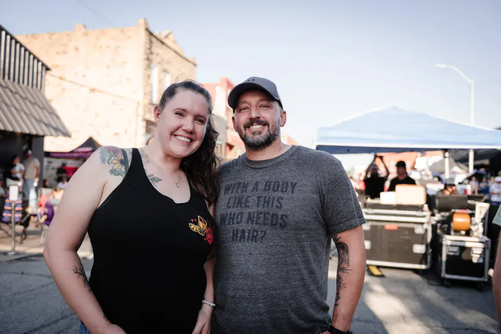 Two smiling festival-goers pose together at Dancing Rabbit Music Festival with venue structures and crowds visible behind them.