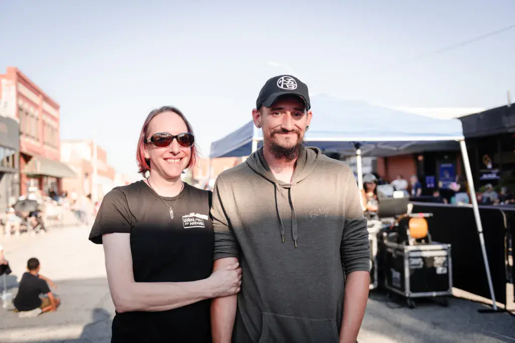Two people pose together at an outdoor music festival venue with buildings and festival infrastructure visible behind them.