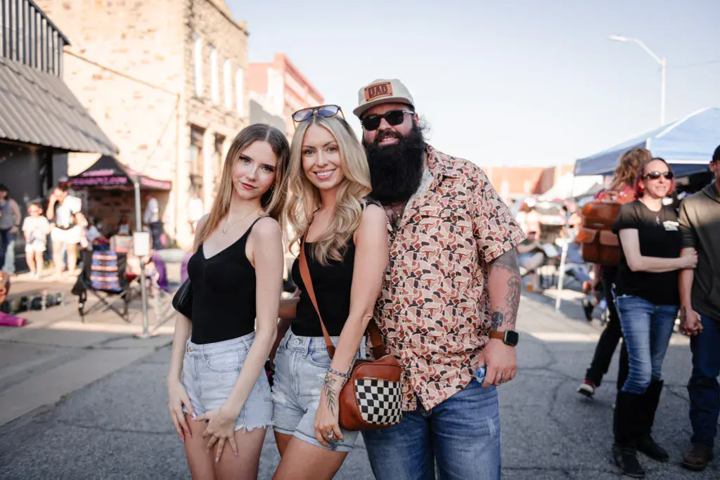 Three festival attendees posing together in front of historic buildings at an outdoor music festival.