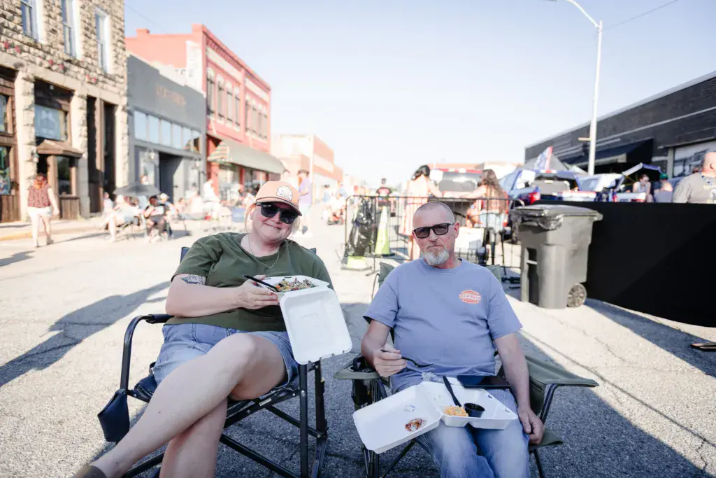 Two attendees enjoy food and drinks while seated in chairs at the Dancing Rabbit Music Festival venue.