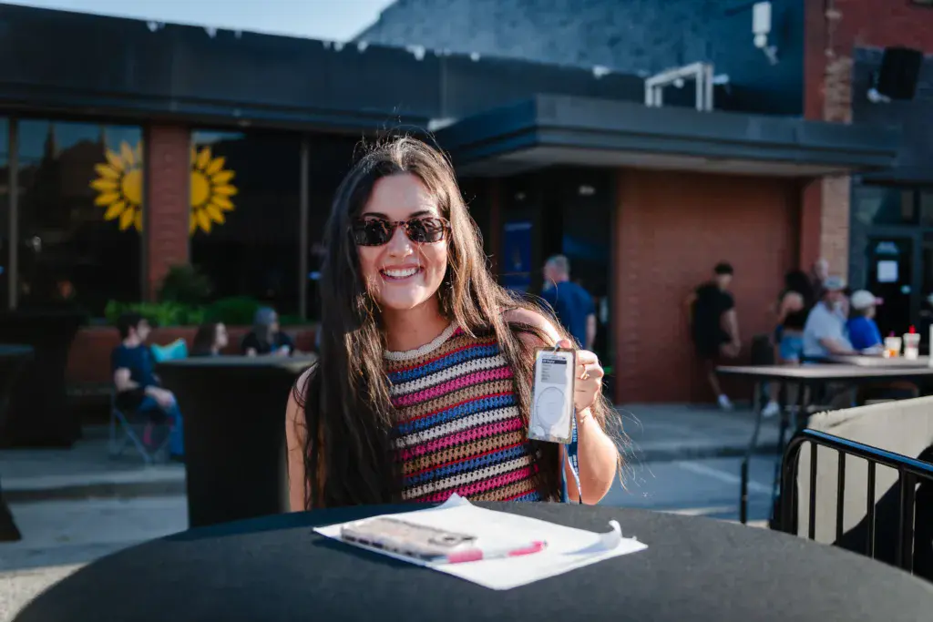 A smiling person wearing sunglasses and a colorful striped shirt holds a festival wristband at an outdoor venue table.