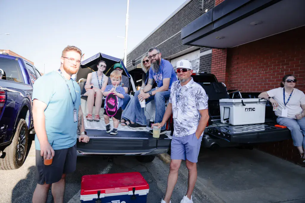 Festival attendees with badges gather around vehicle tailgates and coolers at an outdoor event near a brick building.