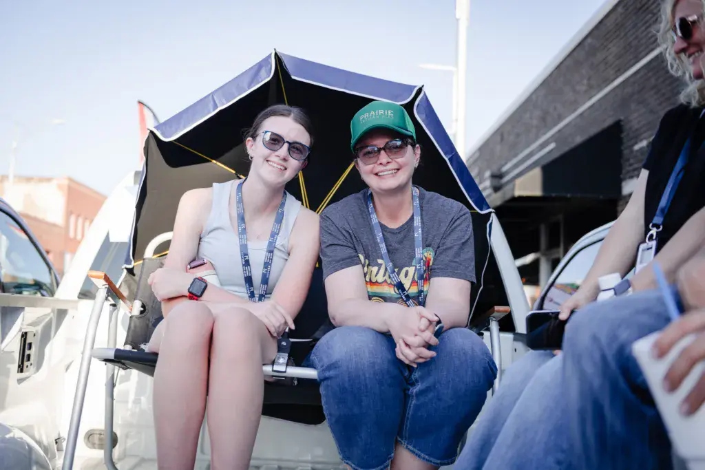 Two festival attendees wearing lanyards smile while sitting under a black umbrella in a truck bed.