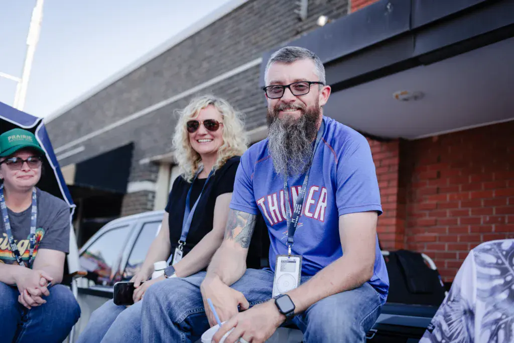 Three festival attendees wearing lanyards smile while seated together against a brick building backdrop.