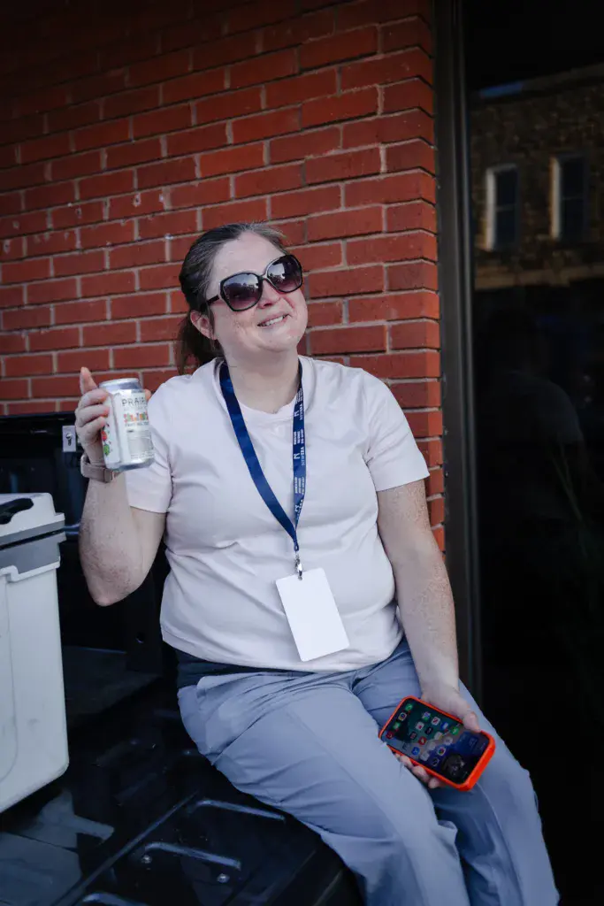 A festival volunteer wearing sunglasses and a lanyard holds a beverage while sitting against a red brick wall.