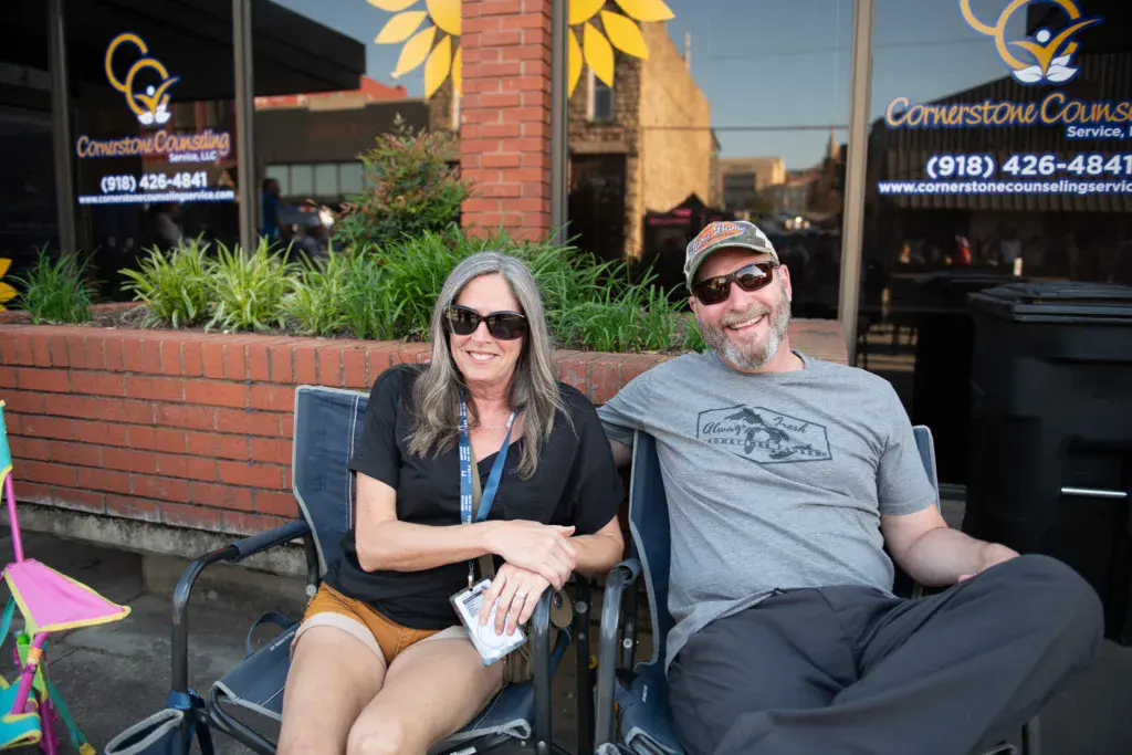 Two people sitting in folding chairs at an outdoor festival venue in front of a brick building with business signage.