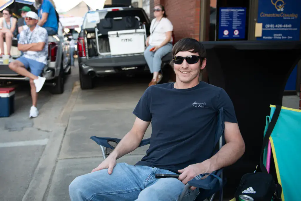 A person wearing sunglasses and a dark t-shirt sits in a chair on a street near parked cars and festival spectators.