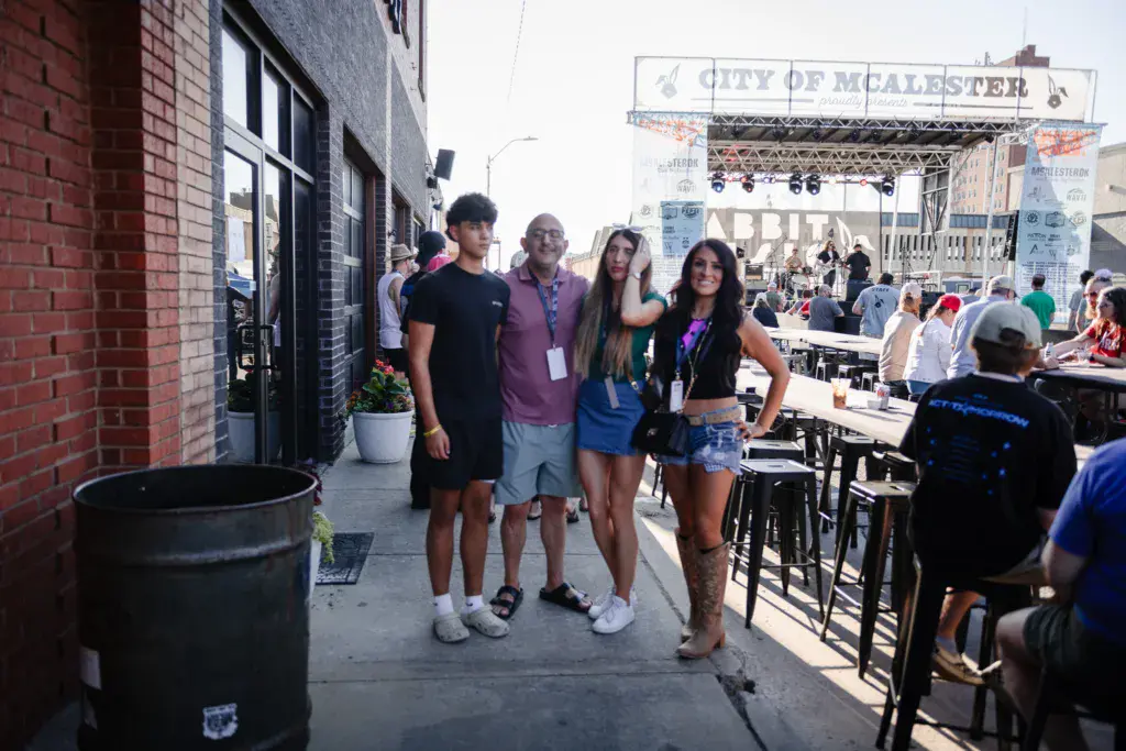 Festival attendees pose together on a brick sidewalk with the main stage and City of McAlester banner visible in the background.