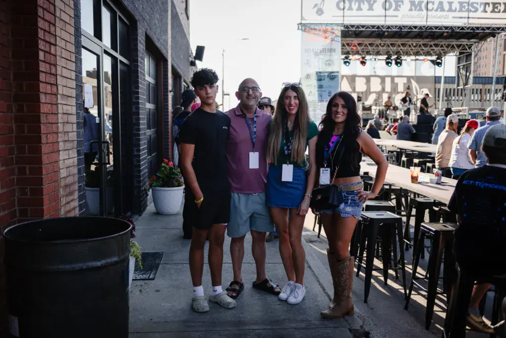 Four people wearing festival credentials stand together on a patio, with the stage and crowd visible behind them.