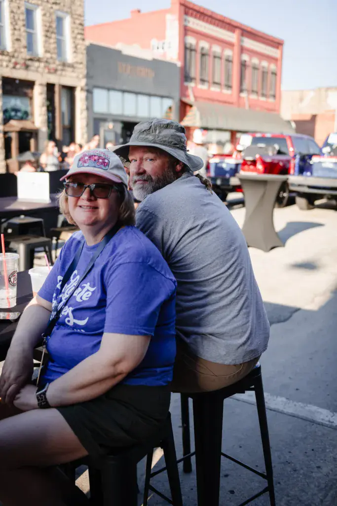 Two festival attendees smile at the camera while seated outdoors in front of historic downtown buildings.