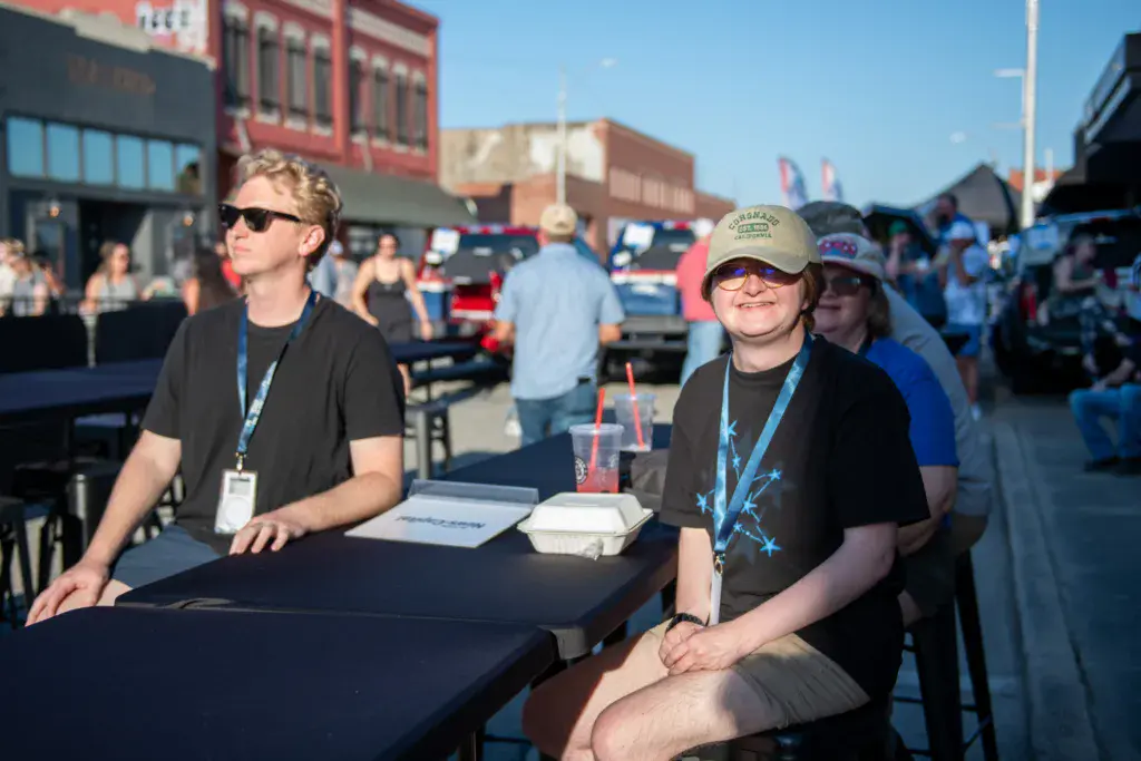 Two festival staff members wearing lanyards sit at a table in an outdoor venue with crowds and historic brick buildings in the background.