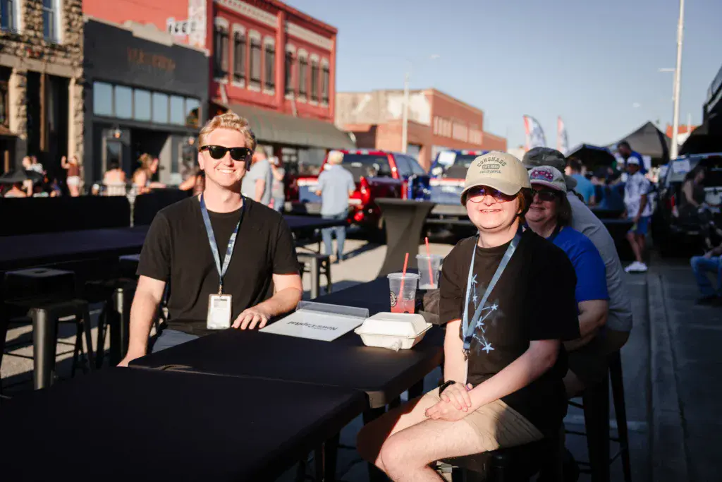 Two festival staff members wearing lanyards sit at a table in an outdoor venue with historic brick buildings and crowds in the background.