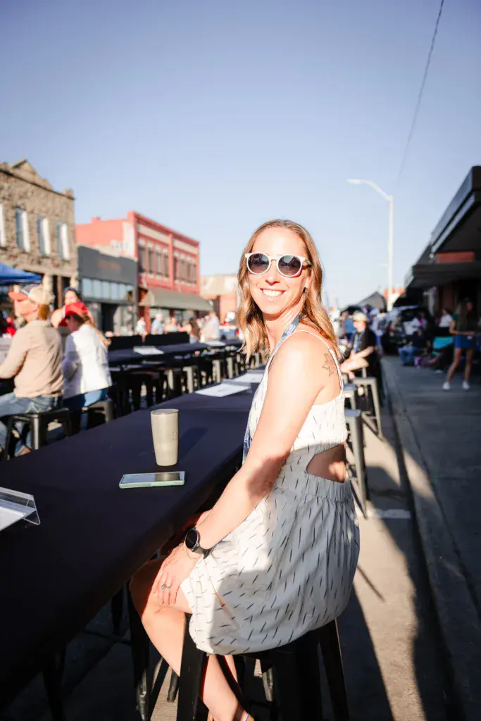 A woman in sunglasses and light-colored outfit stands at an outdoor festival venue with crowd and buildings in background.