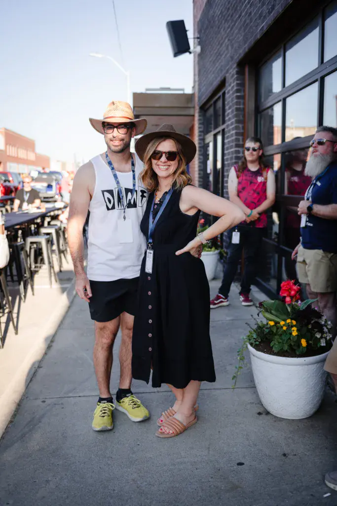 Two festival attendees pose together on a brick sidewalk near decorated planters and other guests.