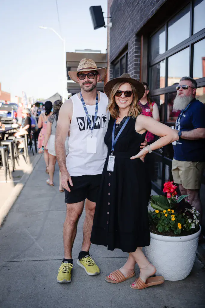 Two festival attendees wearing lanyards pose together on a sidewalk next to a brick building.