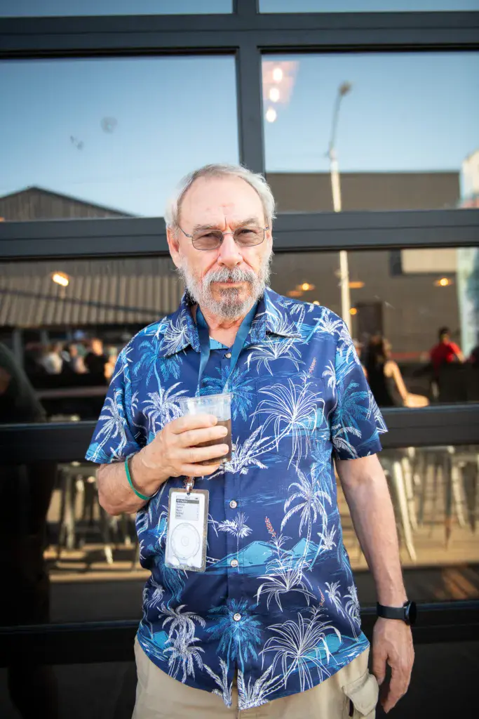 A man wearing glasses and a blue floral shirt holds a drink while wearing a festival badge at an outdoor venue.