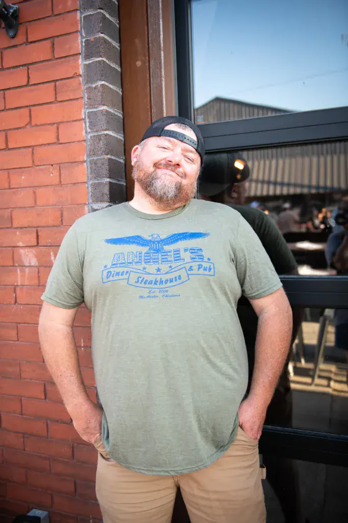 A man in a navy cap and green graphic t-shirt smiles at the camera outside a brick building at the festival.