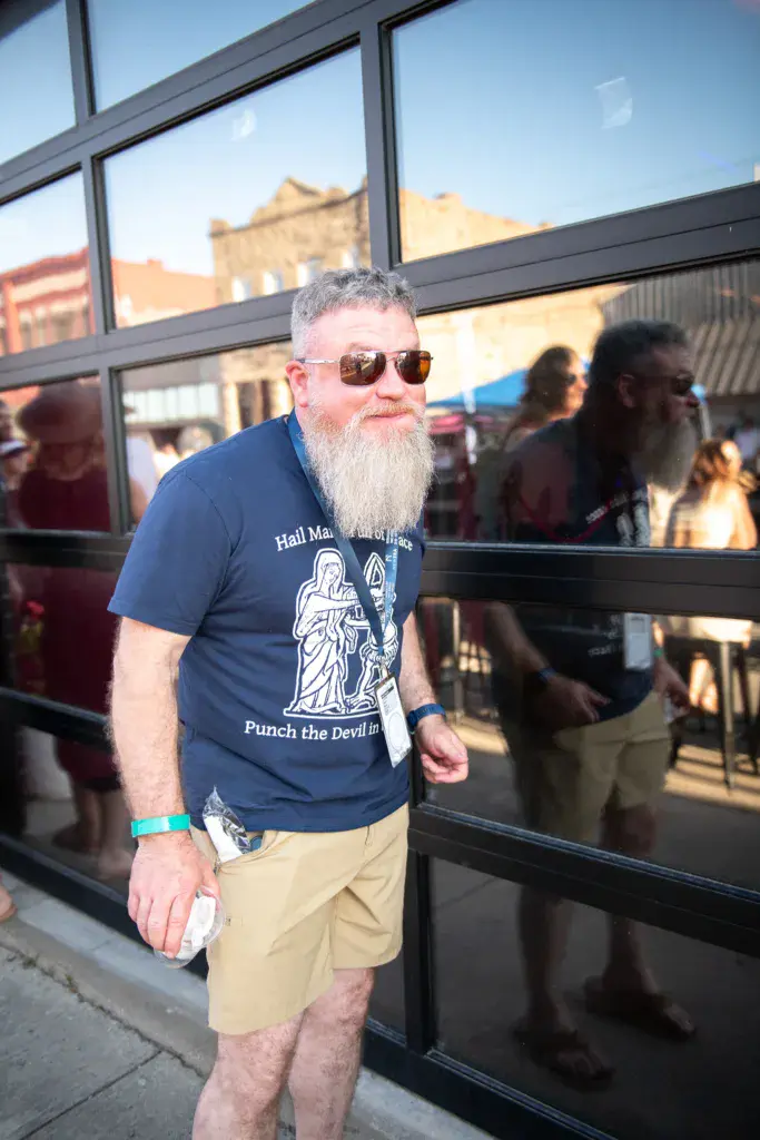 A man with a gray beard wearing sunglasses and a navy graphic t-shirt stands outside a venue at the festival.