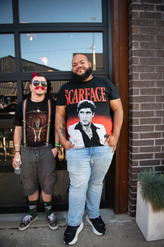 Two festival attendees pose together in front of a brick venue, one wearing a novelty mustache and sunglasses, the other in a Scarface graphic tee.