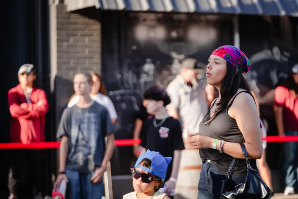 A woman wearing a colorful pink and purple beanie performs or speaks at an outdoor music festival while attendees watch from behind red barriers.