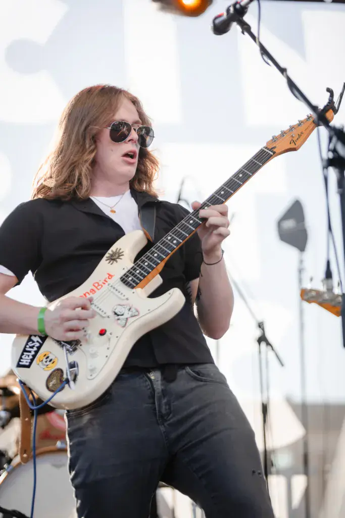 A guitarist with long hair and sunglasses performs on stage with a white electric guitar decorated with patches.