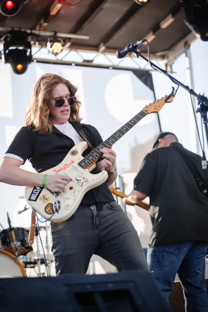 A guitarist wearing sunglasses performs on stage at an outdoor music festival under a covered tent.