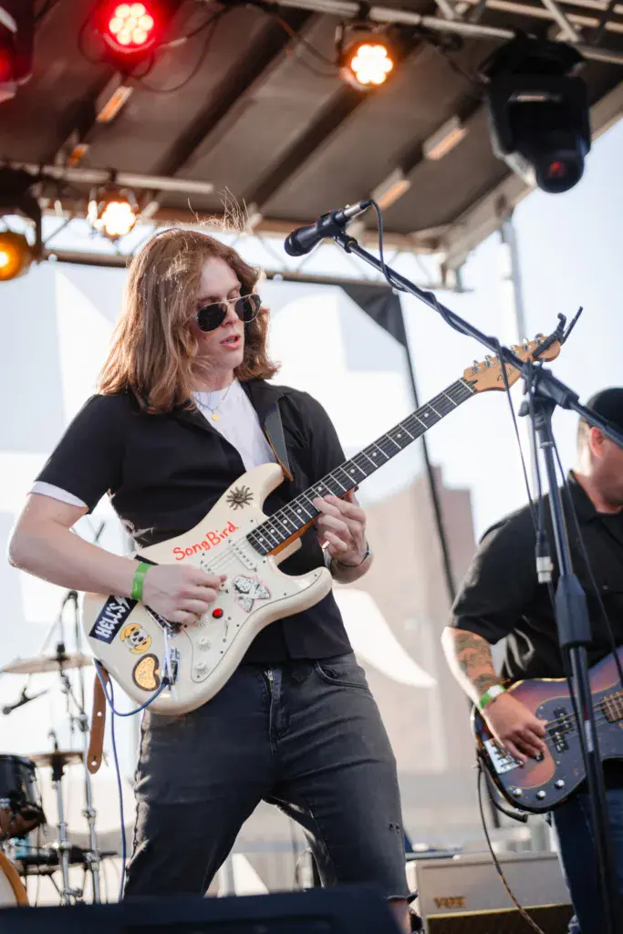 A guitarist in sunglasses and black clothing performs on stage with a decorated white electric guitar.