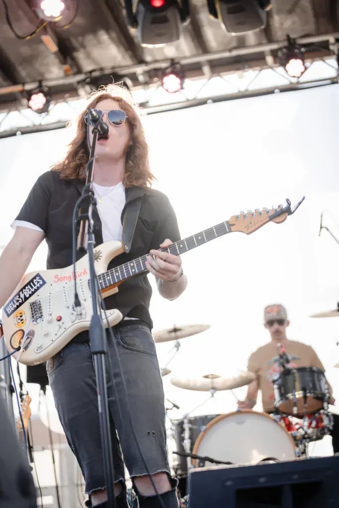 Guitarist and vocalist performing on stage with drummer visible in background under professional lighting.