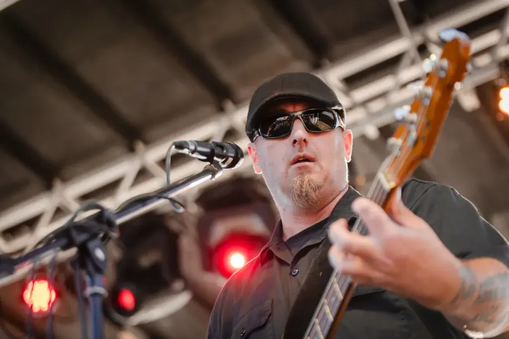 A performer wearing sunglasses and a cap sings into a microphone while playing an electric bass guitar on stage.