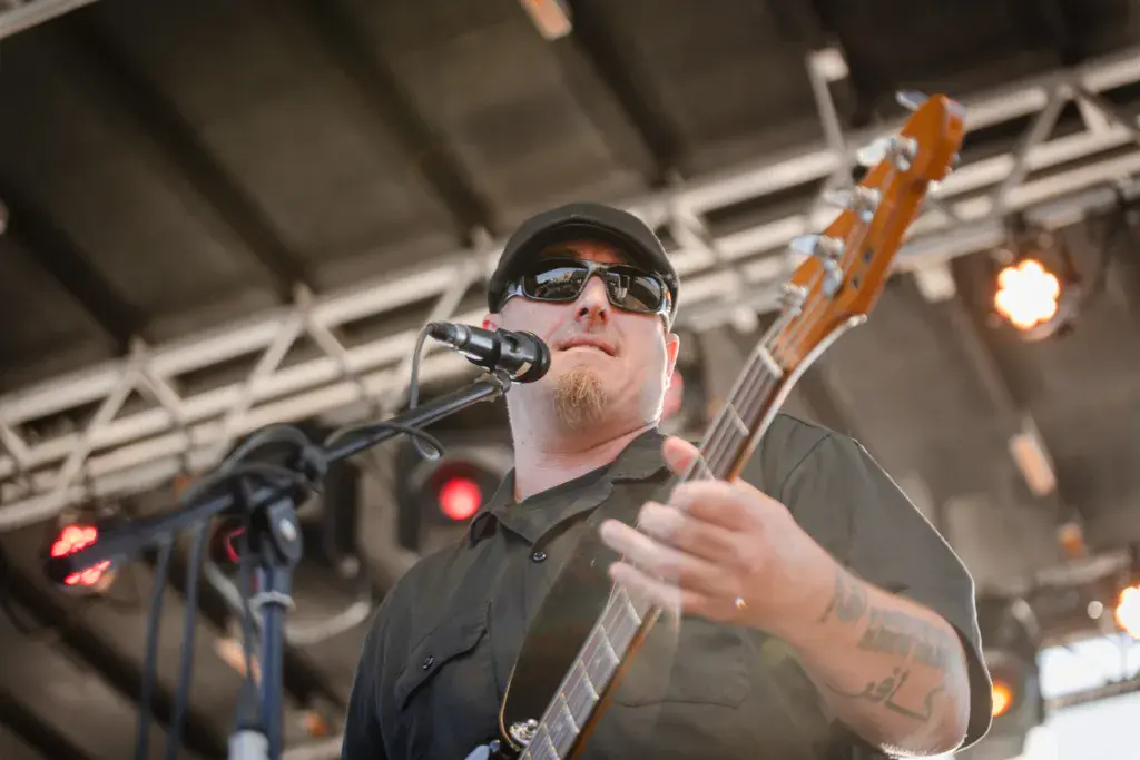 A musician wearing sunglasses and a cap sings into a microphone while playing an acoustic guitar on stage.
