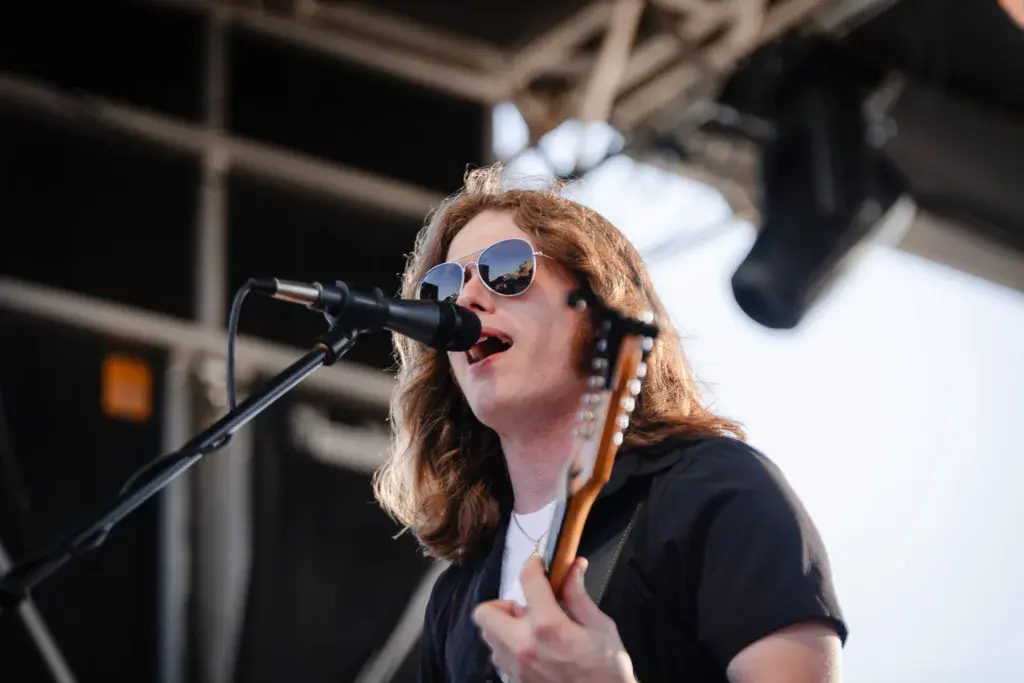 A performer with reddish-brown hair sings into a microphone while holding a guitar on an outdoor stage.