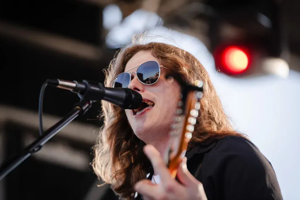 A performer singing into a microphone while wearing sunglasses and holding a guitar on stage.
