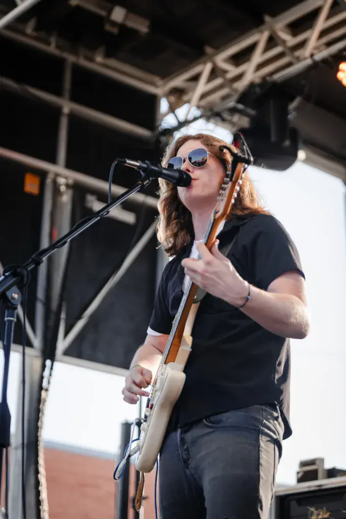 A performer wearing sunglasses and a white headband plays guitar and sings into a microphone on stage.