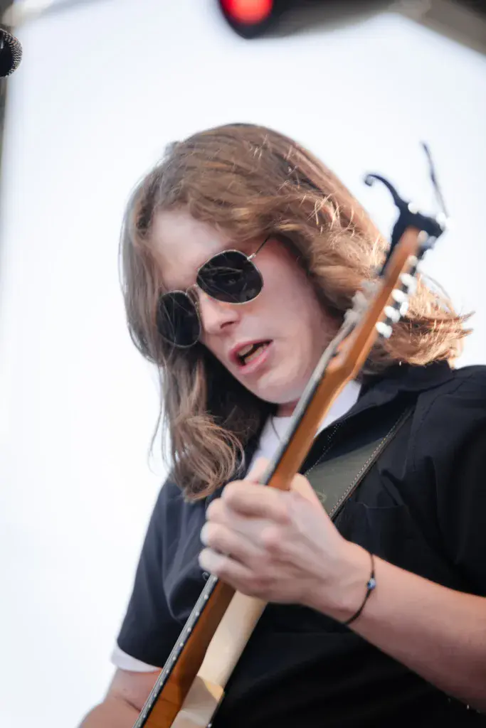 A performer wearing round sunglasses holds a wooden guitar against a white background.