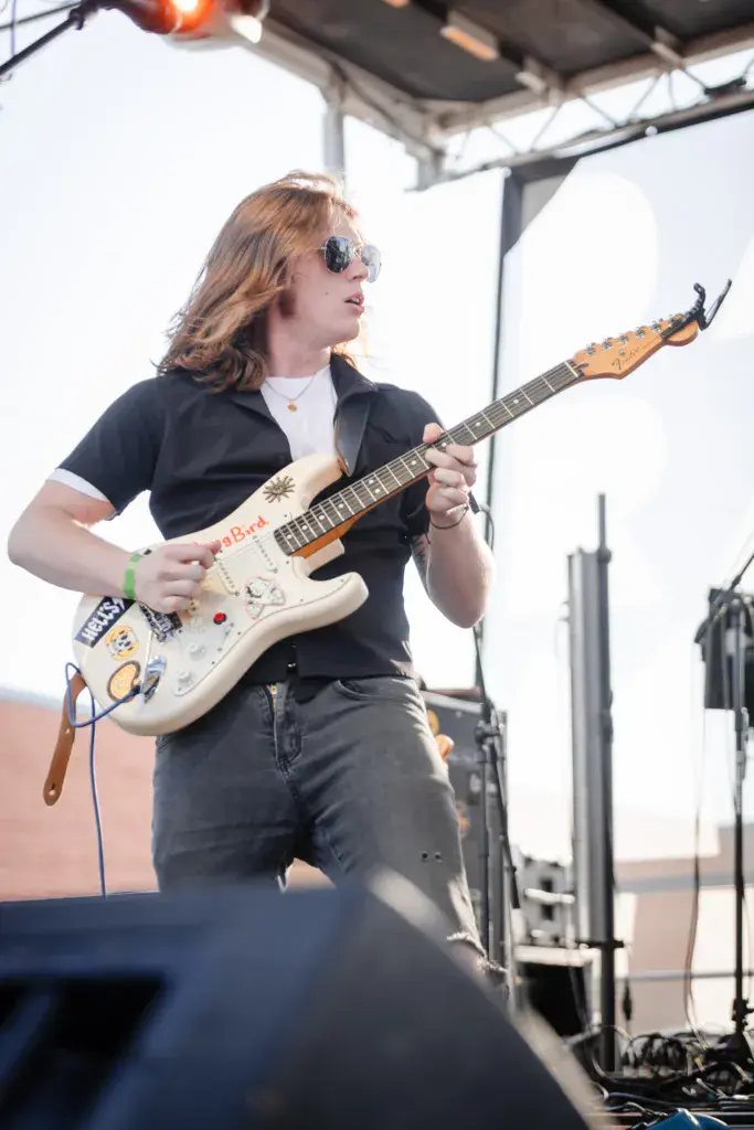 A performer wearing sunglasses and a black shirt plays an electric guitar on stage under a tent structure.