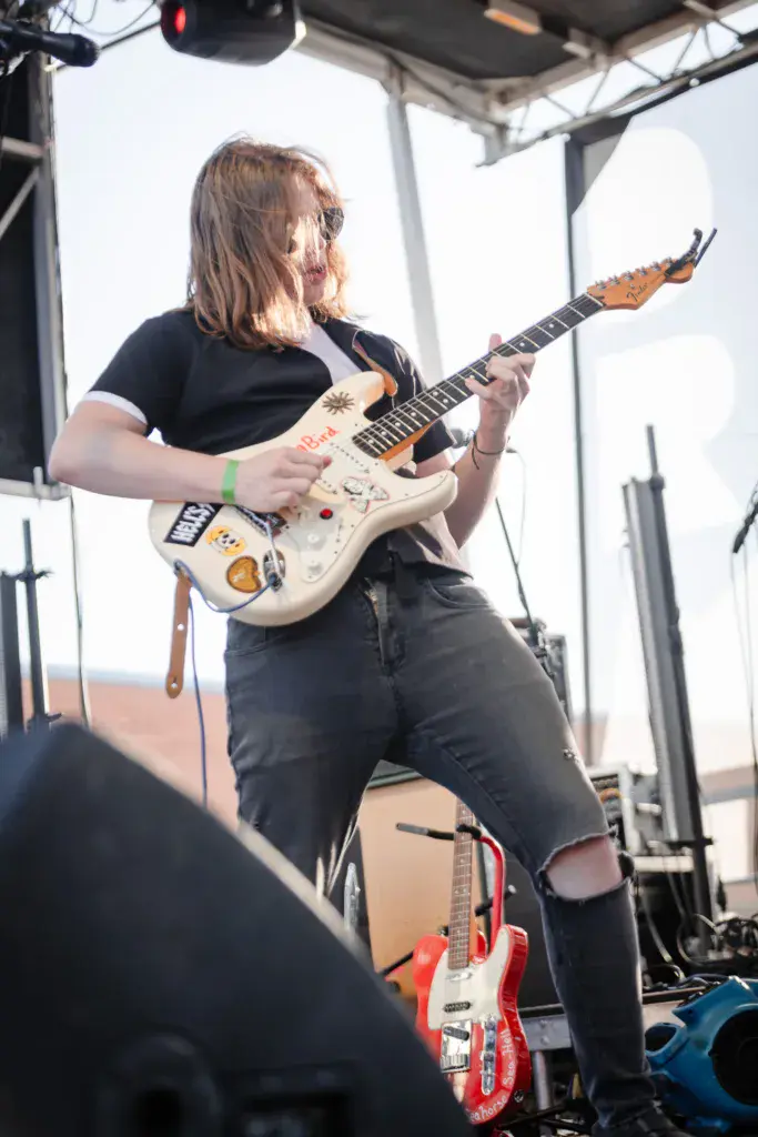 Guitarist with long hair performs on stage under a tent canopy, playing a cream-colored electric guitar.