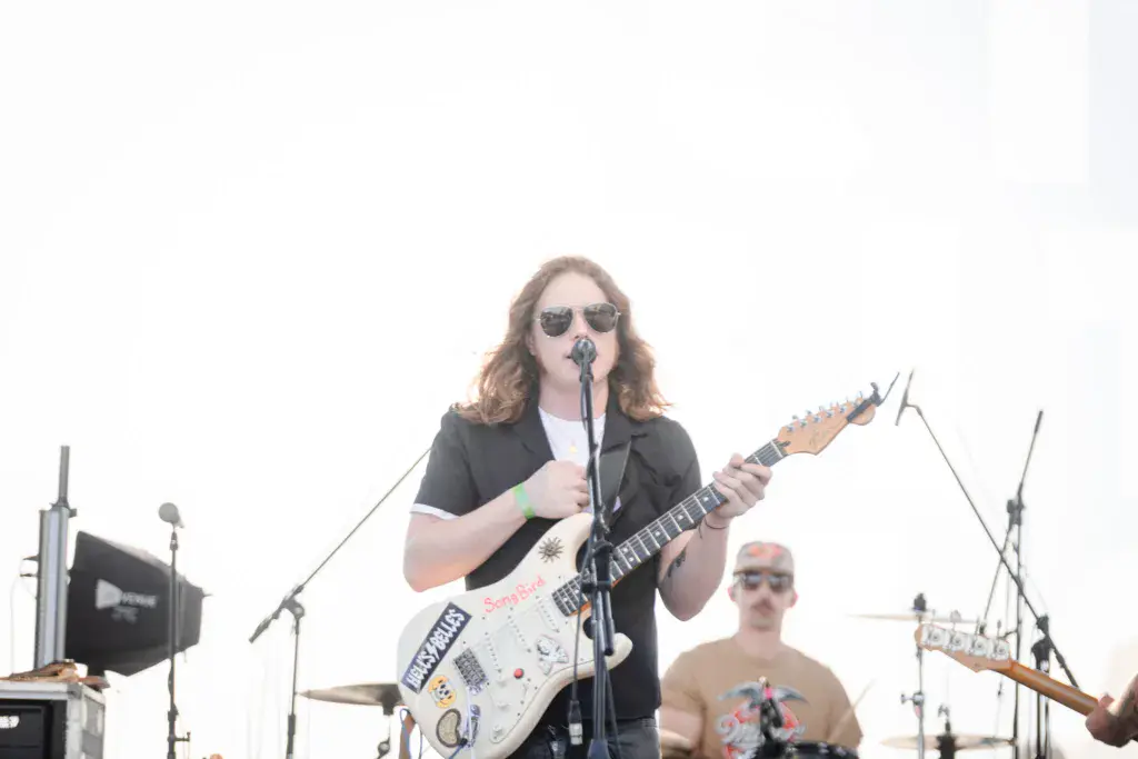 A guitarist wearing sunglasses performs at a stage microphone with band members visible behind.