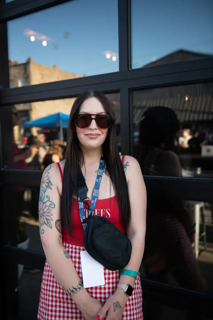 A person wearing sunglasses and a red and white gingham skirt stands at a festival with venue buildings and crowd in the background.