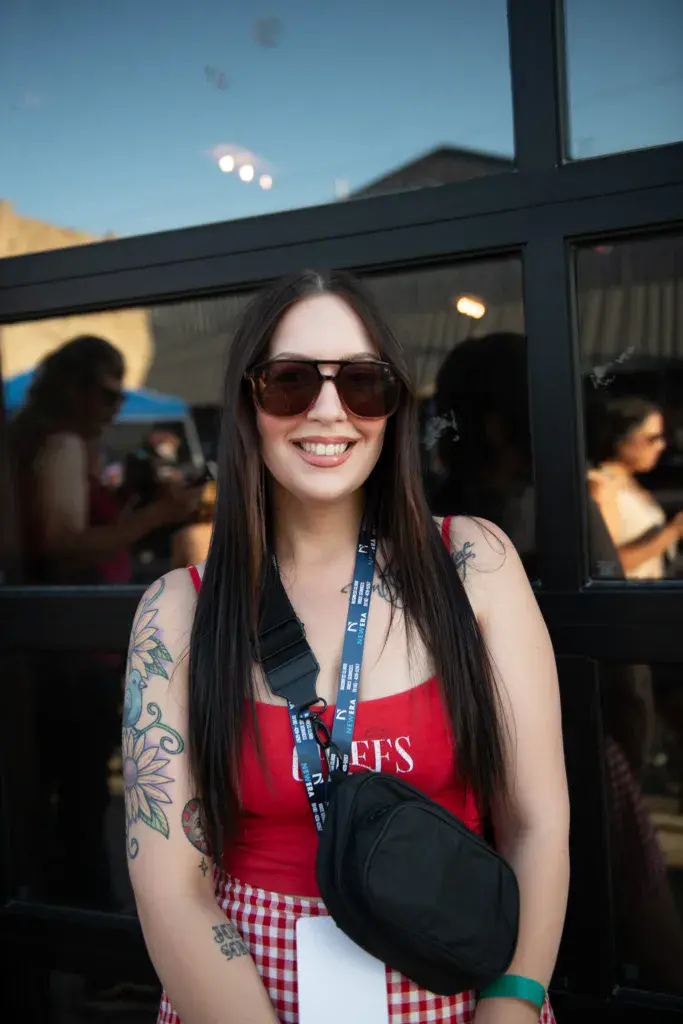 Festival attendee with long dark hair and sunglasses smiling at the camera while wearing a lanyard and red outfit.